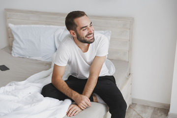 Calm young handsome bearded man sitting in bed with white sheet pillow blanket in bedroom at home....