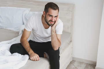 Fototapeta premium Calm young handsome bearded man sitting in bed with white sheet pillow blanket in bedroom at home. Sad upset male spending time in room. Rest relax good mood lifestyle concept. Mock up copy space.