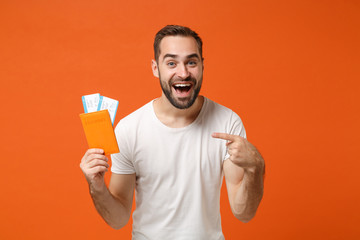 Cheerful young man in casual white t-shirt posing isolated on orange background studio portrait. People lifestyle concept. Mock up copy space. Pointing index finger on passport boarding pass ticket. - obrazy, fototapety, plakaty