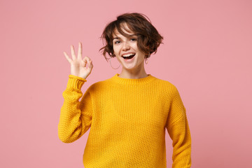 Cheerful young brunette woman girl in yellow sweater posing isolated on pastel pink wall background, studio portrait. People sincere emotions lifestyle concept. Mock up copy space. Showing OK gesture.
