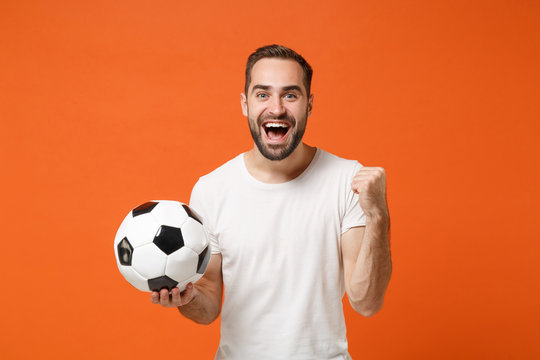 Excited young man in casual white t-shirt posing isolated on orange background studio portrait. People sincere emotions lifestyle concept. Mock up copy space. Hold soccer ball doing winner gesture.