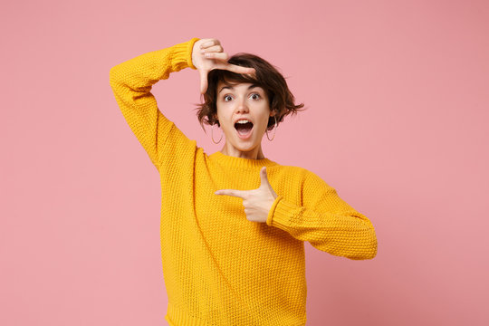 Shocked young brunette woman girl in yellow sweater posing isolated on pink background in studio. People lifestyle concept. Mock up copy space. Keeping mouth open, making hands photo frame gesture.