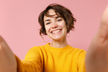 Close up of smiling young brunette woman girl in yellow sweater posing isolated on pastel pink background in studio. People lifestyle concept. Mock up copy space. Doing selfie shot on mobile phone.