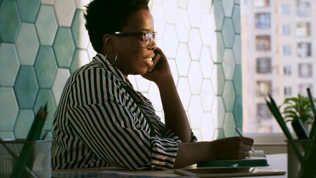 PAN shot of smiling african american businesswoman in eyeglasses writing on notepad and speaking on mobile phone in the office