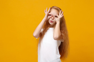 Funny little ginger kid girl 12-13 years old in white t-shirt isolated on yellow background. Childhood lifestyle concept. Mock up copy space. Holding hands near eyes, imitating binoculars or glasses.