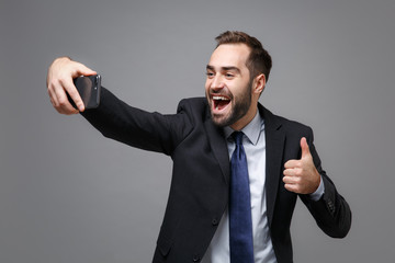 Cheerful young business man in suit shirt tie posing isolated on grey background. Achievement career wealth business concept. Mock up copy space. Doing selfie shot on mobile phone, showing thumb up.