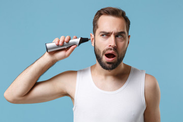 Close up bearded young man 20s years old in white shirt hold electric trimmer isolated on blue...