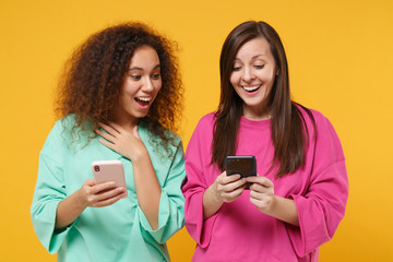 Fototapeta premium Two excited women friends european african american girls in pink green clothes posing isolated on yellow background. People lifestyle concept. Mock up copy space. Using mobile phone, typing message.
