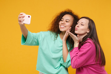 Two women friends european and african girls in pink green clothes posing isolated on yellow background. People lifestyle concept. Mock up copy space. Doing selfie shot on mobile phone, send air kiss.