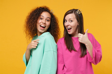 Fototapeta na wymiar Two women friends european african american girls in pink green clothes, crowns posing isolated on yellow background. People lifestyle concept. Mock up copy space. Point index finger showing thumb up.
