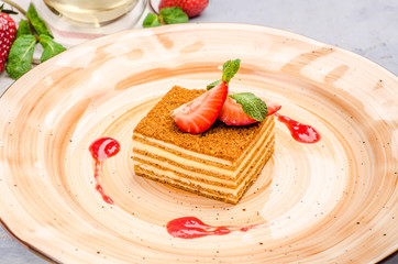 Delicious piece of honey cake served with fresh summer berry on a plate, close up on a background teapot. Homemade russian Medovik decorated with mint and strawberry.
