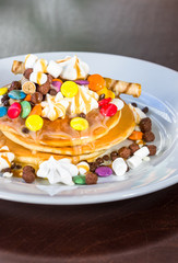Pancakes in a white plate are decorated with colorful candies, marshmallows and topped with caramel topping