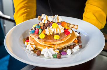 Pancakes in a white plate are decorated with colorful candies, marshmallows and topped with caramel topping. The plate is held in hands