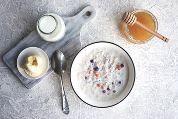  Bowl of oatmeal porridge with berries, honey, bottle of milk, butter, wooden rustic board, grey table near window, hot and healthy food for Breakfast, top view, flat lay.           