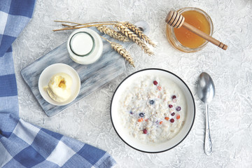  Bowl of oatmeal porridge with berries, honey, bottle of milk, butter, wooden rustic board, grey table near window, hot and healthy food for Breakfast, top view, flat lay.           