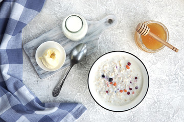  Bowl of oatmeal porridge with berries, honey, bottle of milk, butter, wooden rustic board, grey table near window, hot and healthy food for Breakfast, top view, flat lay.           