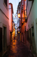 Lisbon, Portugal - July, 24th, 2018 : Night falls on a picturesque alley of the Alfama, the oldest district of Lisbon. Santo Estevao bell tower in background.