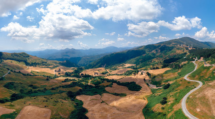 Fototapeta premium Aerial wide view panorama of fields and winding roads, farms, harvested grass, haystacks. Wind turbines in mountain, Passo Di Cento, Taglieto, Varese Ligure, Province of La Spezia, Italy