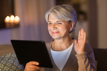 technology, old age and people concept - happy senior woman with tablet pc computer having video call and waving hand at home in evening