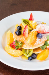Pancakes on a white plate, decorated with fruits: strawberries, mangoes, orange, kiwi and blueberries and drizzled with natural honey and sprinkled with powdered sugar.Pancakes on a white plate, decor