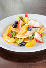 Pancakes on a white plate, decorated with fruits: strawberries, mangoes, orange, kiwi and blueberries and drizzled with natural honey and sprinkled with powdered sugar.Pancakes on a white plate, decor