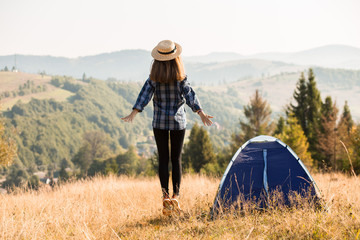 Young beautiful woman in blue shirt and straw hat near tent at autumn landscape background with mountains at horizon