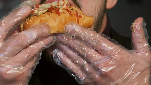 Extra close-up young man bites a hot dog in transparent gloves