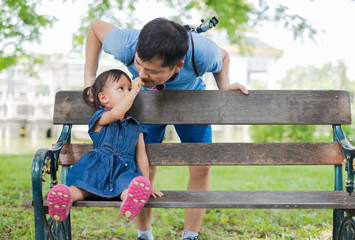 Fototapeta premium Cute asian little girl sitting on the wooden chair in the public park and she gives some piece of cookies for her father, concept of love in family life, daughter and father.