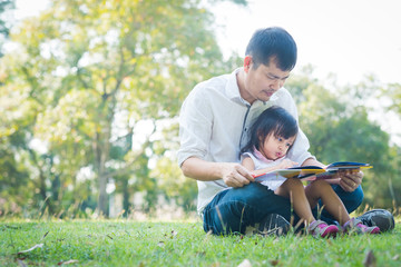 Asian father and daughter are reading the book together in the park with fully happiness moment, concept of learning activity for kid in family lifestyle.