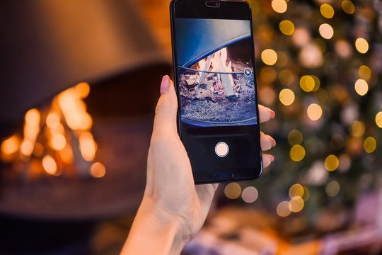 female hand with manicure holding a smartphone and taking a photo of fire in the fireplace with the background of a Christmas tree in the lights