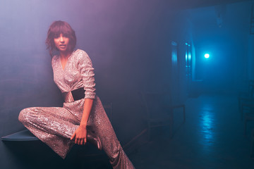 dark-haired girl sitting on a table in blue and violet light in a silver jumpsuit with a wide belt and cowboy shoes, she has silver big earrings, professional makeup and looks into the camera