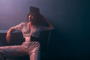 dark-haired girl sitting on a chair in violet light in a silver jumpsuit with a wide belt and...