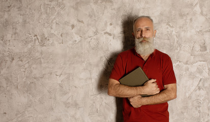 Bearded Senior man stands on grey wall and holding a book.