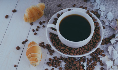 Cup of coffee with coffee beans and a croissant on vintage wooden table. Homemade croissant served with black coffee or americano. Coffee break with delicious croissant. Autumn composition still life.