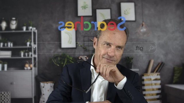 Handsome mature businessman in front of the computer screen at work or at home typing in an anonymous search engine the word GIFT for a festivity. Christmas. View through the pc monitor. Cine lens.