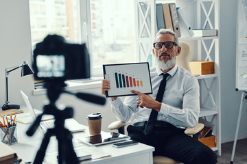 Confident mature man in elegant shirt and tie showing chart while making social media video