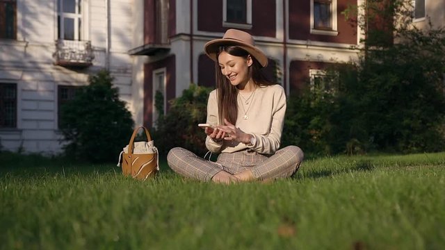 Beautiful portrait of good-looking happy smiling woman in stylish hat which relaxing on the lawn near houses and using her smartphone