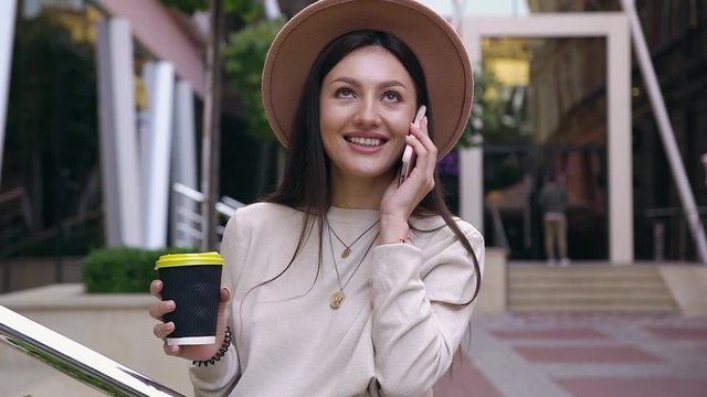 Adorable smiling young woman with long brown hair in stylish hat talking on phone while drinking her coffee near modern office building