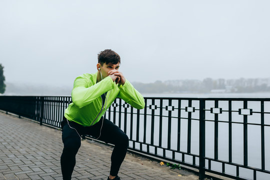 Outdoor image of young athletic man exercising squats at city lake background. Fit male doing squats outdoors in morning in the park on fog background. Sport, lifestyle and people concept.