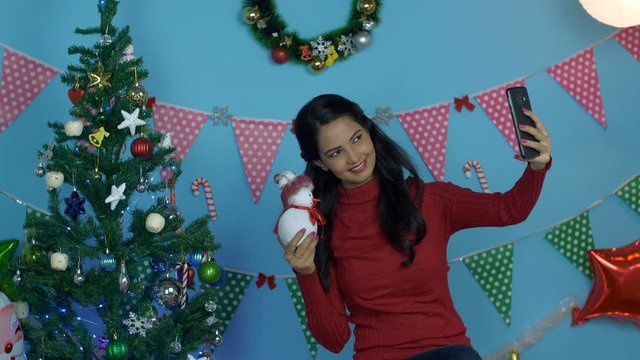 Pretty Indian girl clicking selfies with a small toy snowman during Christmas season. Young woman having fun while taking pictures and decorating the Christmas tree at home - Christmas Eve