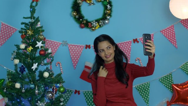 Beautiful young woman having fun while clicking pictures on Christmas Eve - winter season. Pretty Indian female happily taking selfies with a decorated Christmas tree and colorful festive background