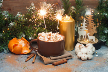 Fototapeta na wymiar Brown cup with marshmallow and burning bengal lights. There are golden candle, tangerine, cinnamon rolls and christmas tree garland around.