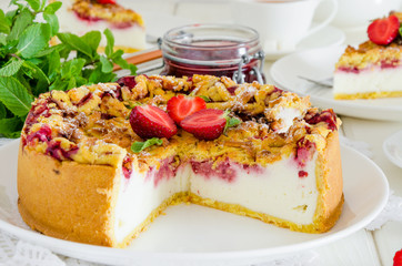 Baked creamy cheesecake with strawberries and crispy shtrezel on a plate on a white wooden background with a cup of tea.
