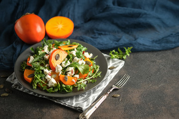 Healthy salad with persimmons, arugula, feta cheese and pumpkin seeds on a dark plate on a dark background. Vegetarian menu, healthy food. Prevention of vitamin deficiency. Top view.