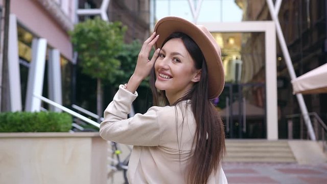Good-looking happy young lady with long brown hair dressed in beautiful blouse and stylish hat looking at camera during her walk near modern building