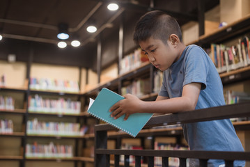 Asian boy student standing and reading a book at the library in the school.Students search for books in the bookshelf.Portrait Asian boy.