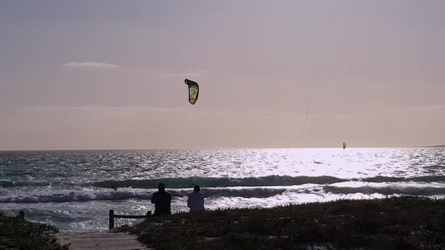 Silhouette people at beach watching kiteboarders catch airtime and jumps. Windy summer sunset evening in Cape Town. Kitesurfing on glistening sea. Silver mirror ocean waves. Holiday in paradise.