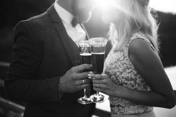 Black and white foto the glasses with champagne in the hands of the bride and groom. Newlyweds clinking glasses and enjoying romantic moment together on a bridge on wedding day. Wedding details.