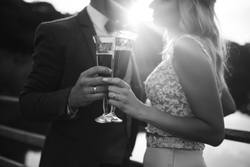 Black and white foto the glasses with champagne in the hands of the bride and groom. Newlyweds clinking glasses and enjoying romantic moment together on a bridge on wedding day. Wedding details.