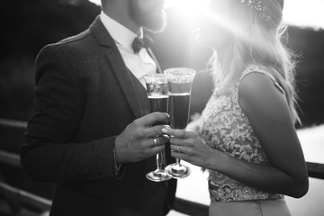 Black and white foto the glasses with champagne in the hands of the bride and groom. Newlyweds clinking glasses and enjoying romantic moment together on a bridge on wedding day. Wedding details.